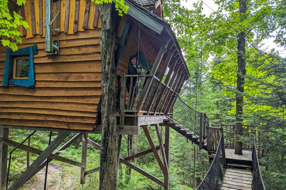 Treehouse accommodation at Les Toits du Monde, with Dharma standing on the balcony and a wooden bridge leading to the entrance, surrounded by dense forest.