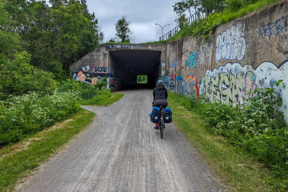 Dharma riding towards a graffiti-covered tunnel along the P'tit Train du Nord trail, with greenery on both sides.