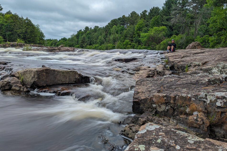 Dharma sitting on rocks next to a rushing river with whitewater rapids, surrounded by lush forest.