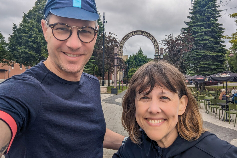 Dharma and Devon posing for a selfie in front of the entrance arch to Parc Linéaire Le P'tit Train du Nord, with outdoor seating and trees in the background.