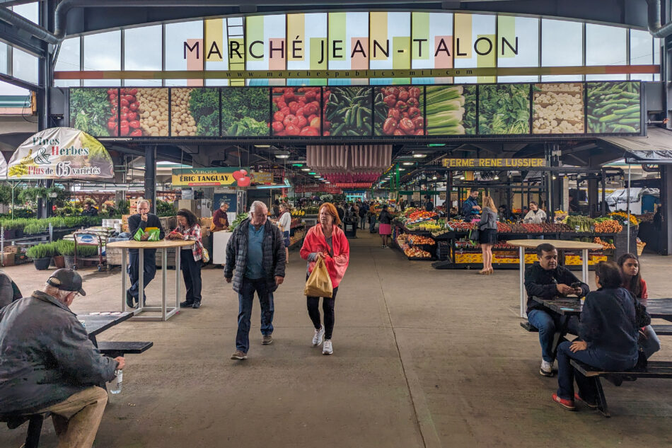 Busy scene at Marché Jean-Talon in Montreal, with shoppers walking past vibrant fruit and vegetable stalls under a large sign featuring images of fresh produce.