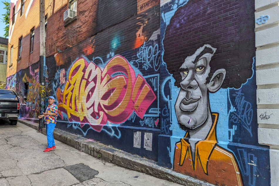 Vibrant street mural featuring a caricature of a man in a suit and colorful abstract designs on a city alley wall, with a person posing in a patterned outfit in the foreground.