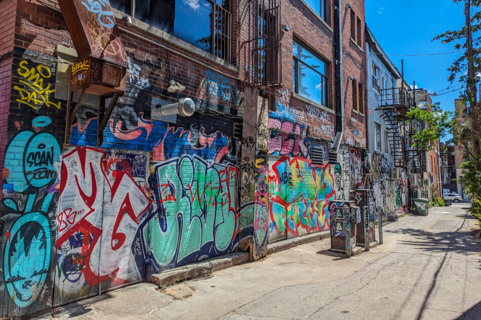 Urban street scene showcasing buildings covered with various graffiti and murals, including colorful abstract patterns and character art, in a vibrant, artistic neighborhood.