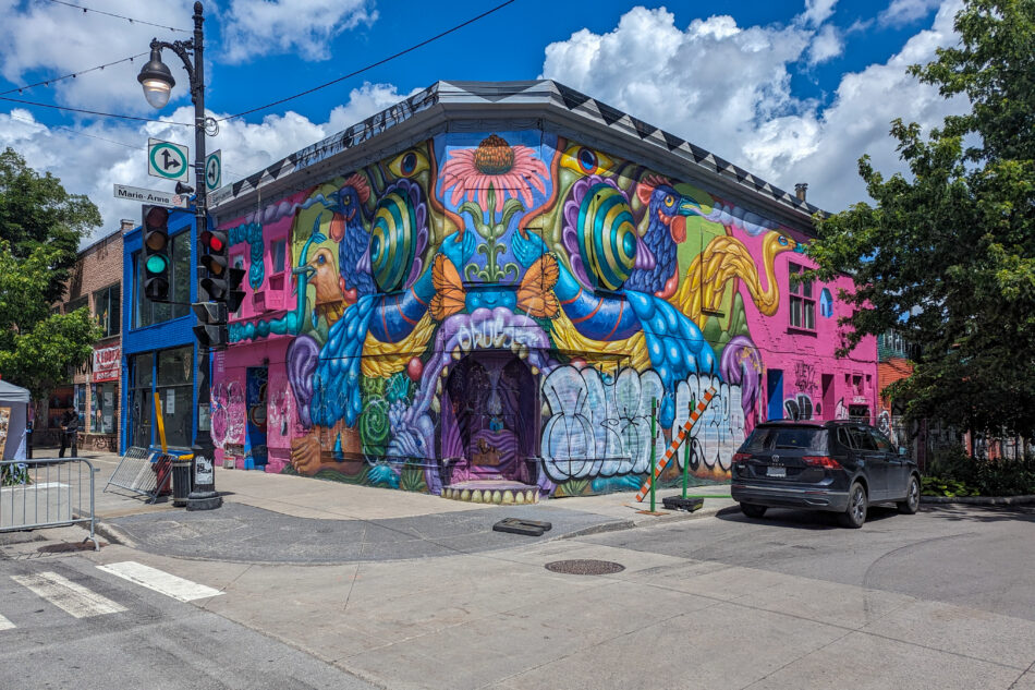 Bright and colorful mural covering the entire facade of a corner building, depicting fantastical creatures and floral patterns in a lively urban setting.