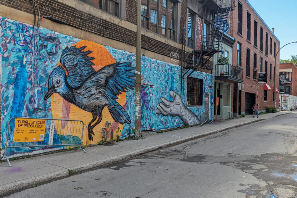 Striking mural of a blue bird in motion, painted on the side of an urban building, surrounded by abstract blue patterns and a sketched human hand, creating a dynamic street art scene.