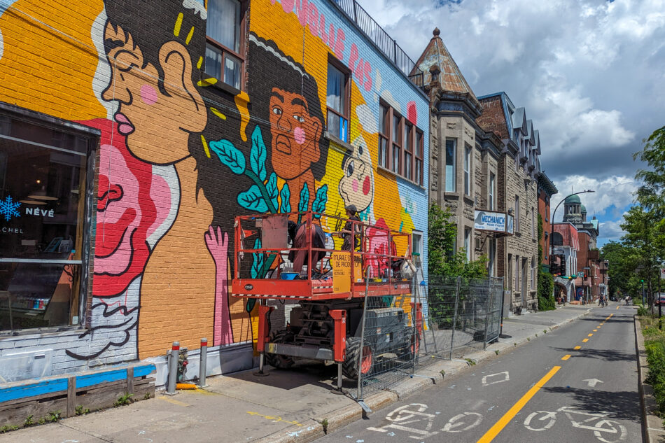 Wall mural featuring large cartoon figures painted in bold colors on a yellow background, with ongoing work by an artist on a lift, adding vibrant life to an urban street.