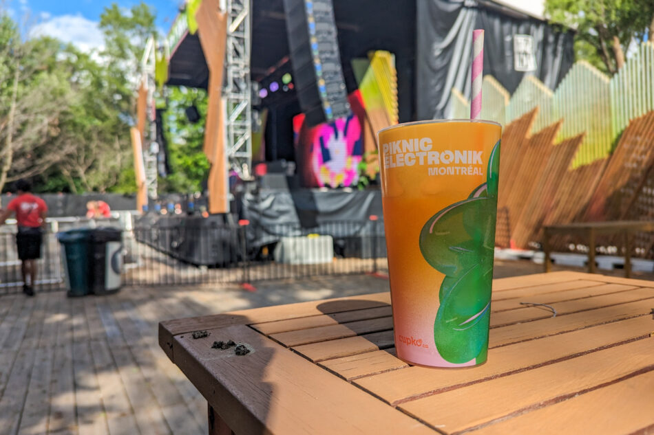 Close-up of a colorful drink with a pink straw in a Piknic Electronik branded cup, placed on a wooden table with a blurred festival stage in the background.