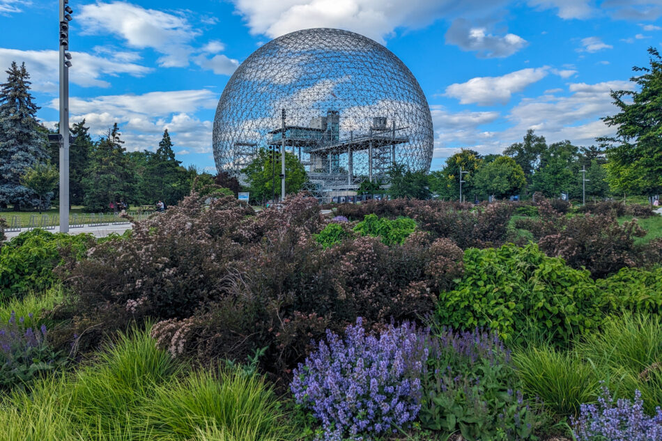 The Montreal Biosphere, a geodesic dome, viewed across lush green and purple flower beds under a bright blue sky, symbolizing a blend of nature and science.