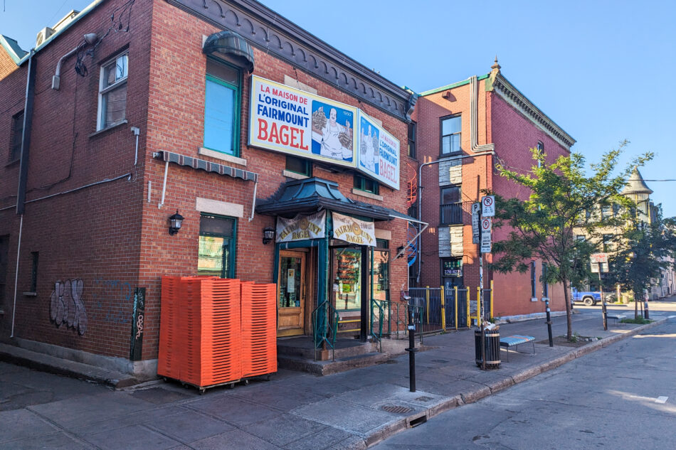 Exterior view of La Maison de l'Original Fairmount Bagel in Montreal, a renowned bakery set in a traditional red brick building, with colorful banners promoting their famous bagels, on a sunny street corner.