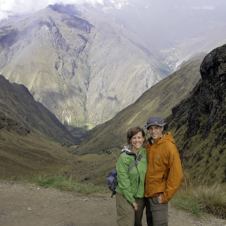 Dharma (left) and Devon (right) in hiking gear standing on a mountain trail with dramatic, fog-covered peaks and valleys in the background, during a trek in Peru.