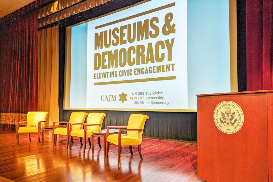 Photograph of the stage in the McGowan Theater at the National Archives Museum with the title of the conference projected onto a screen in the background.
