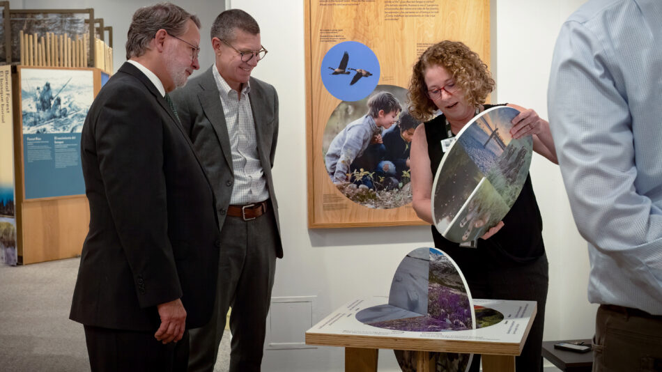 Alt text: Senator Gary Peters (D-MI) is on the far left, wearing a dark suit, white shirt, and tie, attentively observing an interactive exhibit. Next to him, a man in a gray suit and glasses is also watching. A woman with curly hair and glasses, wearing a black sleeveless top, is demonstrating the exhibit, which consists of rotating circular panels displaying images of nature and wildlife. Another person in a light blue shirt is partially visible on the right. The background features educational panels with images of animals, birds, and a child exploring nature, suggesting a conservation or environmental theme.