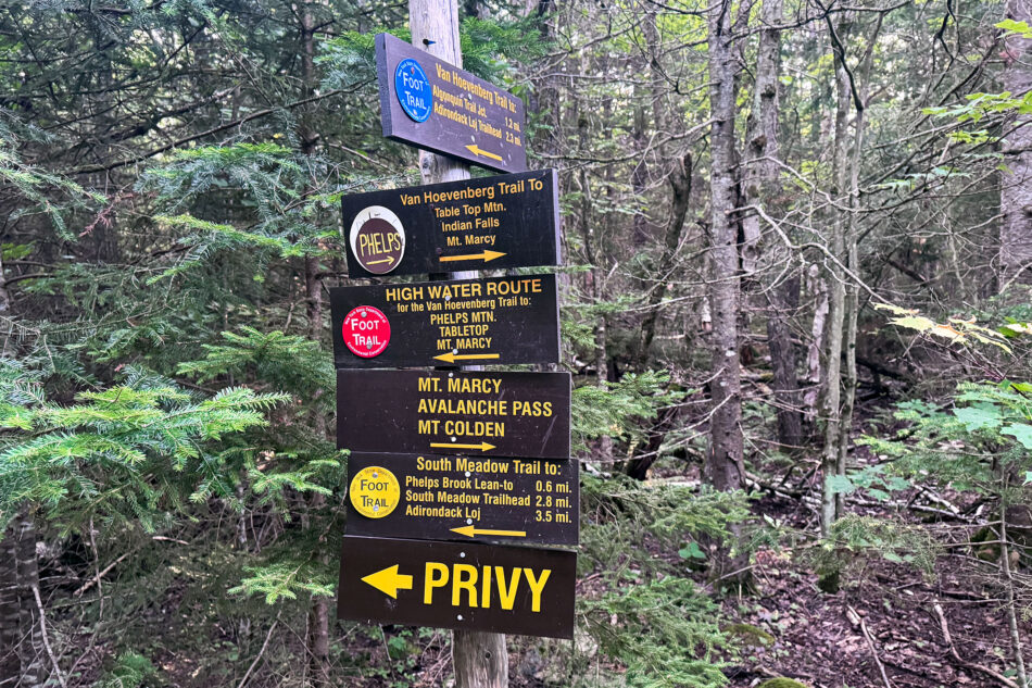 A trail signpost in the woods with multiple wooden directional signs, including routes to Table Top Mountain, Phelps Mountain, Mount Marcy, Avalanche Pass, and Mount Colden, plus a sign pointing to a privy.