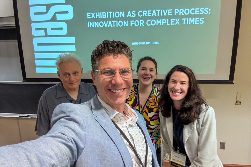 Devon Akmon takes a selfie with MSU Museum CoLab Studio colleagues Mark Sullivan, Caroline White, and Abbie Stevens in front of a presentation screen displaying "Exhibition as Creative Process: Innovation for Complex Times" at the AAMG annual conference.